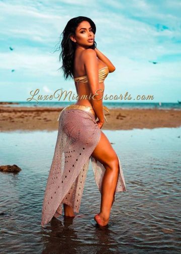Brunette escort Miranda posing on a beach in golden swimwear and white throw on