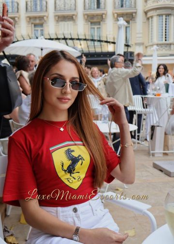 Brunette Miami escort Kathy sitting at the outdoor restaurant, wearing red Ferrari shirt and white pants