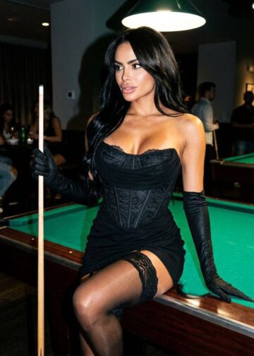 Brunette Latina escort model Miranda posing by the billiard table, wearing short black dress and black stallings