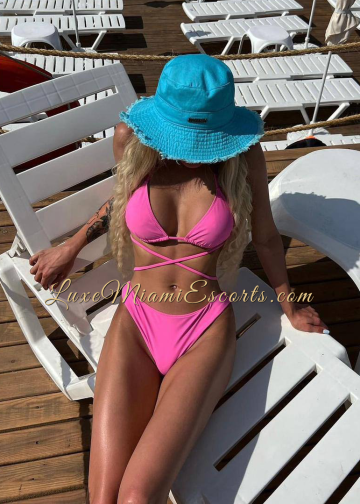 Dina posing on pool chair, wearing pink swimwear and blue hat. Amateur photo.